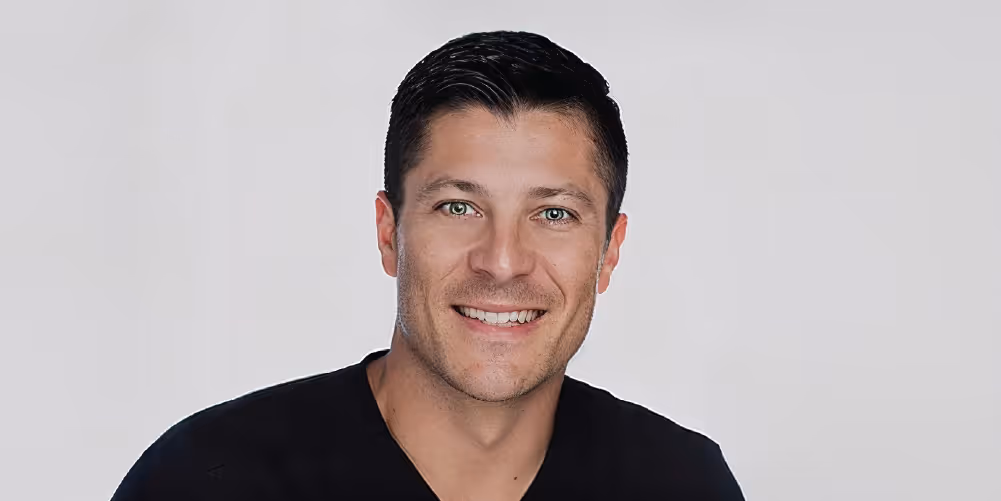 A smiling man with short black hair and green eyes wearing a black scrub top, showcasing a friendly appearance.