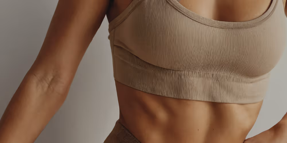 A close-up image of a woman's torso wearing a beige crop top, showing defined abdominal muscles and a well-toned physique.