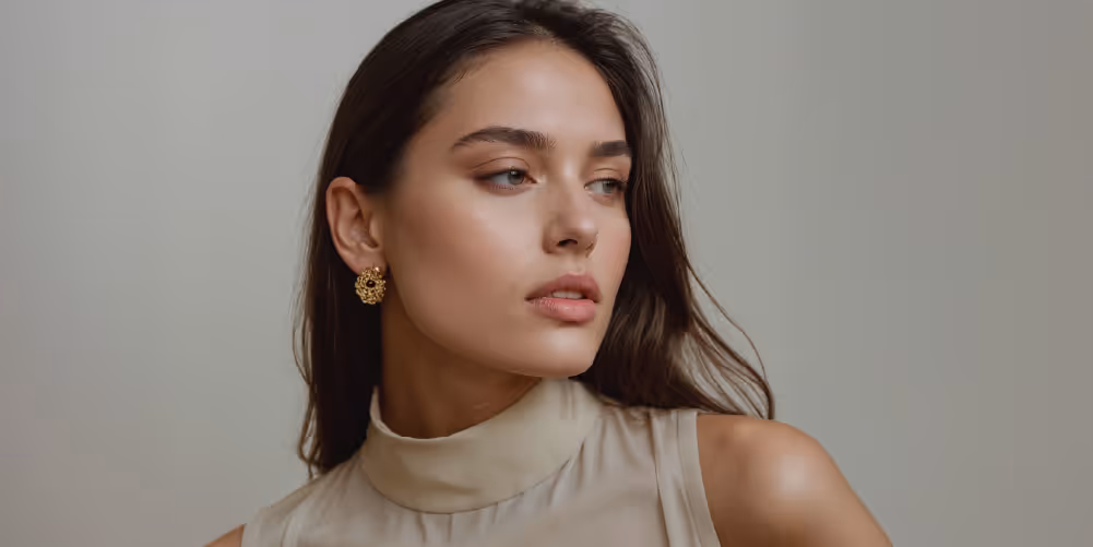 A woman with long, dark hair wearing a sleeveless beige top and gold stud earrings, looking to her right.